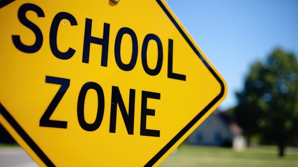 Yellow school zone traffic sign with blurred trees and houses in the background.