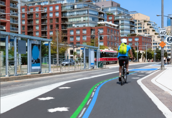 A cyclist in a helmet and bright yellow backpack rides in a dedicated, separated bicycle lane marked with blue and green lines next to a street and transit stop in Toronto, Canada.