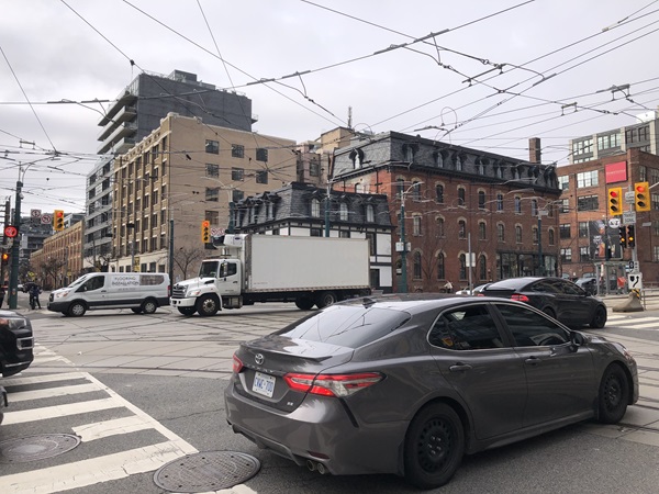 A busy Toronto street intersection featuring a gray sedan in the foreground, with a white delivery truck and a service van navigating a crossing equipped with overhead streetcar power lines and surrounded by multi-story brick buildings.